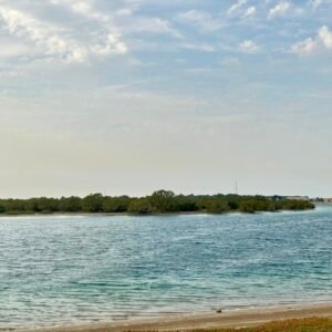 Calm waters at Al Bahya Hidden Beach, with a scenic backdrop of sand, greenery, and a peaceful sky.