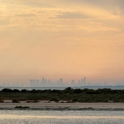 A serene view of Abu Dhabi’s skyline at sunset, seen from a hidden beach with calm waters and lush greenery in the foreground, creating a peaceful and picturesque atmosphere.