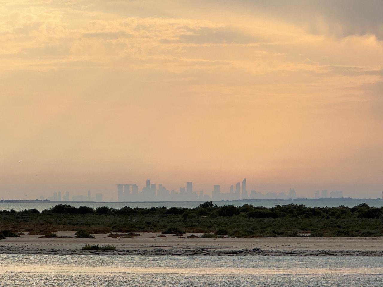A serene view of Abu Dhabi’s skyline at sunset, seen from a hidden beach with calm waters and lush greenery in the foreground, creating a peaceful and picturesque atmosphere.