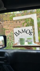 Two cups of iced matcha from Bamo's Café are served on a wooden tray out a car window, with a hand holding the tray in front of a charming outdoor sign that reads "BAMO'S" and colorful handwritten cafe signs in the background.