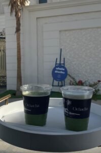 Two cups of iced matcha labeled "Ochacha" are served on a round white tray outdoors in front of a white villa facade and blue Ochacha drive-thru sign in Shakhbout City, Abu Dhabi, during the day. 