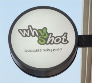 Circular outdoor sign for "whyshot" café, featuring playful green and black lettering and the slogan "because why not?" against a clear blue sky.
